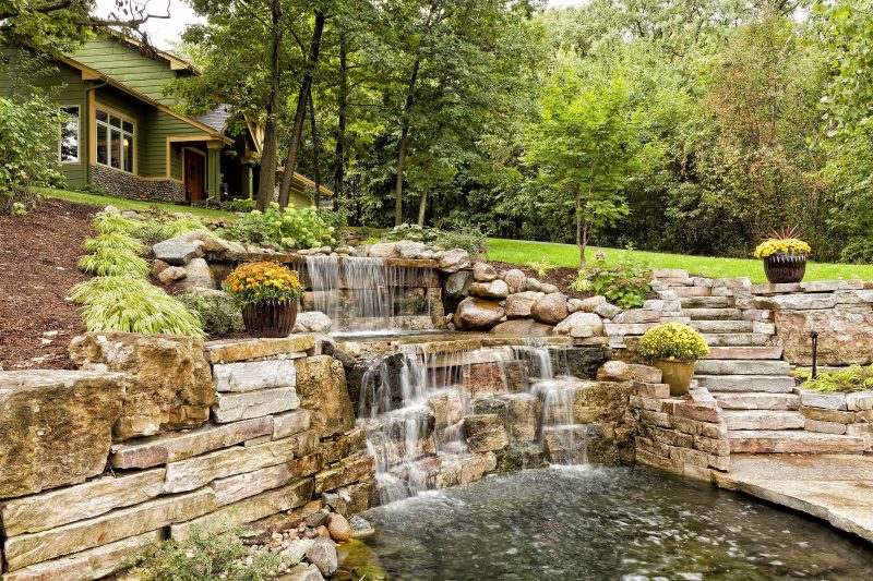 2-level waterfall feature made of river rocks and stone slabs with pond and stairs with same stone slab material and clapboard house siding and stone veneer bottom siding