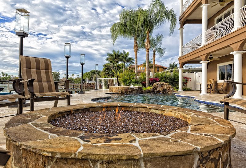 above ground firepit with brown stone veneer and coping with travertine pool surround and patio flooring and pool with blue wave band tiles and lampposts lining the dock