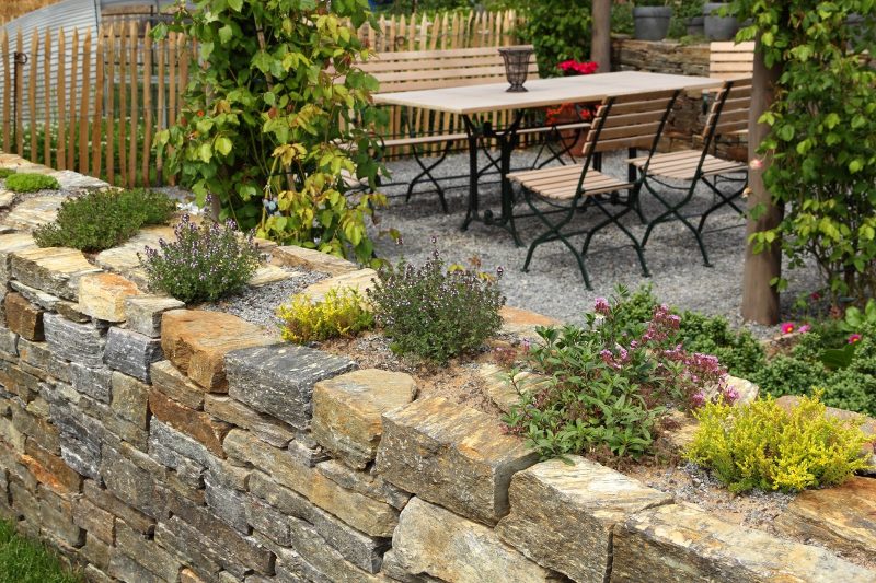 raised beds on stone wall with pea gravel patio flooring and cafe terrace style table and chairs and country picket fence