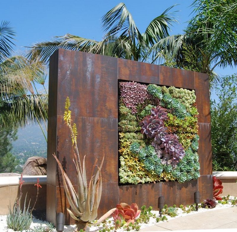 rusted finish vertical custom garden wall with variety in shapes and colors of succulent plants and white pea gravel parterre with floor directional lights and low stucco fence with seating