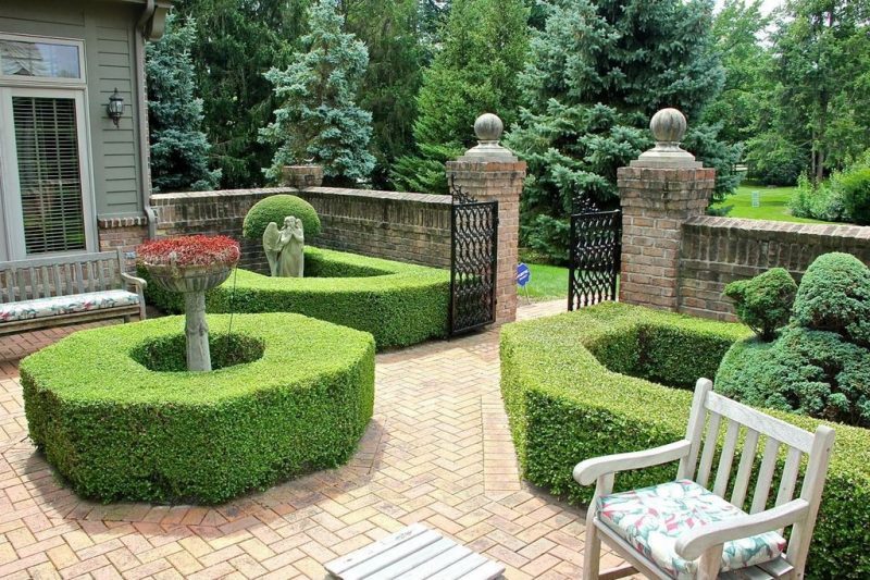 sculpted stone pedestal bird bath with geometric boxwood shrubs and aged brick pave fence and stone angel statue with cast iron garden gate and angel statue