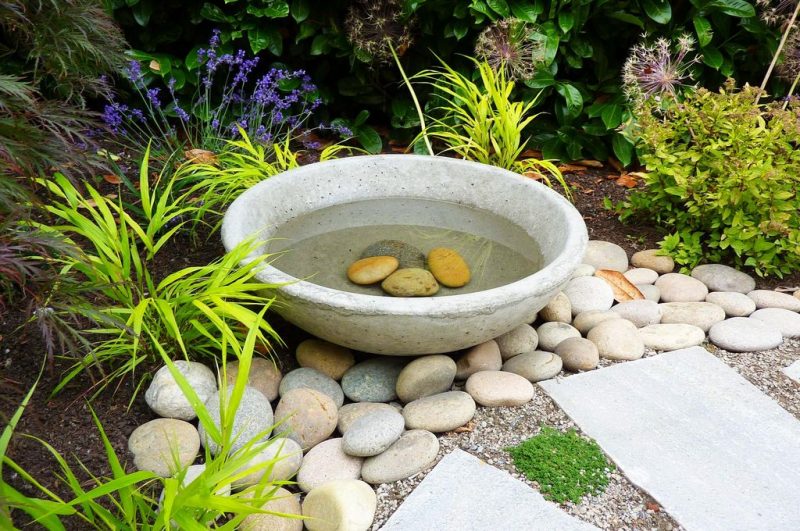 stone ground bird bath with river rocks and rectangular concrete stepping stones with gravel cover around step stones and mulched garden bed