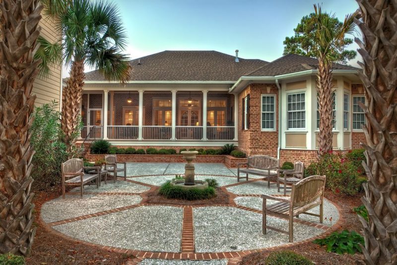 stone pedestal birdbath with water fountain base and pebble aggregate patio flooring and brick inlays with raised brick plant bed and aged wooden benches