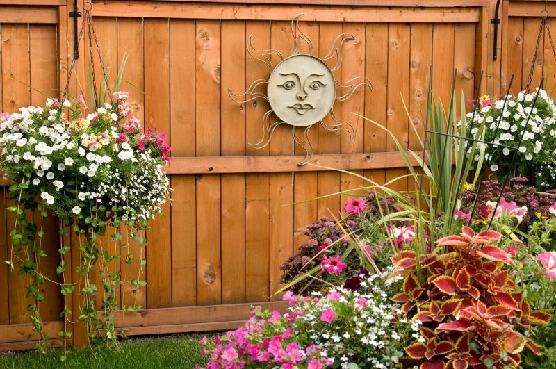 twin planter baskets with white and pink perennials with simple iron basket holder brackets and wooden vertical plank fence and smiley metal sun wall decor with coleus plant