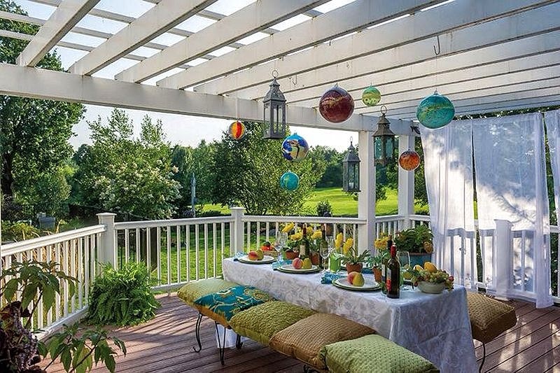 white pergola and purlins with white curtain decor and composite deck floor and extended uncovered patio area and iron bench with cushion seating and decorative balls and lanterns