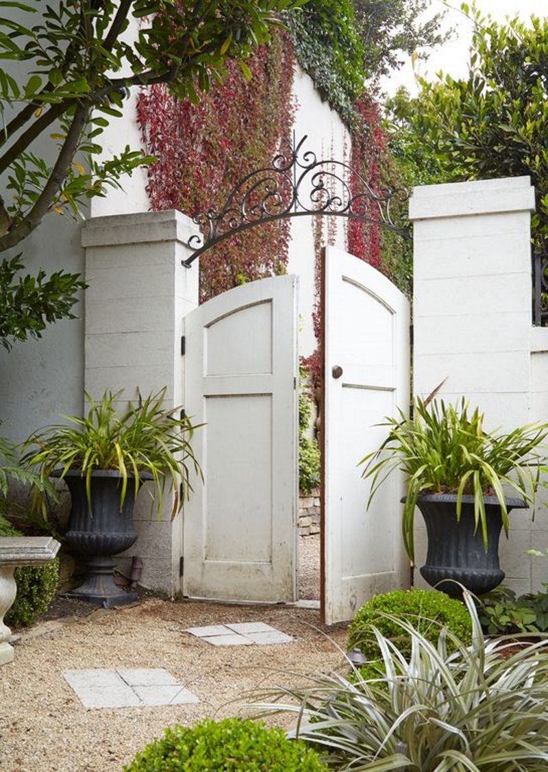 wooden arched gate with wrought iron arbor accent and pair of black urn planters and concrete square stepstone tiles with marble bench