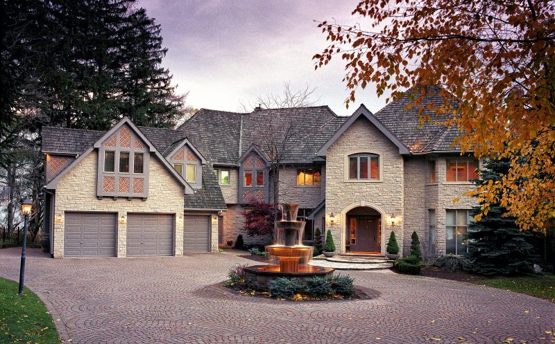 3-tier cascading fountain with stacked stone base and uplighting and circular brick driveway around fountain with red brick and white design pattern on dormer windows and outdoor wall lamps flanking doorway