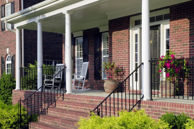 brick siding and porch skirt with running bond pattern and wrought iron railing and stair balustrade calling back shutters and front door with round pillars with roman-inspired capitals and twin antique terracotta planters