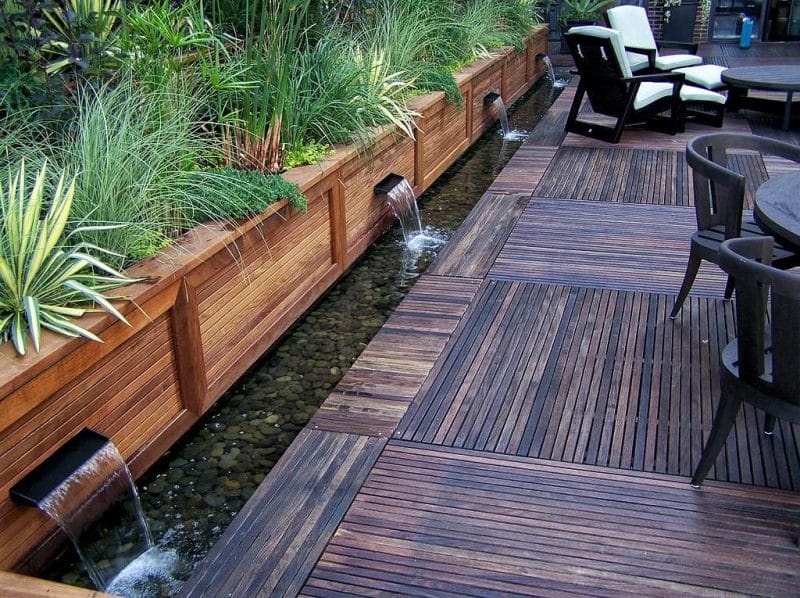 built-in fountains on a stained raised wood garden bed with wood deck tiles and planks and pebbles pond lining and Atlas Fescue and Aureola hakone grass with resin chaise lounge and patio chairs in a dark mahogany finish