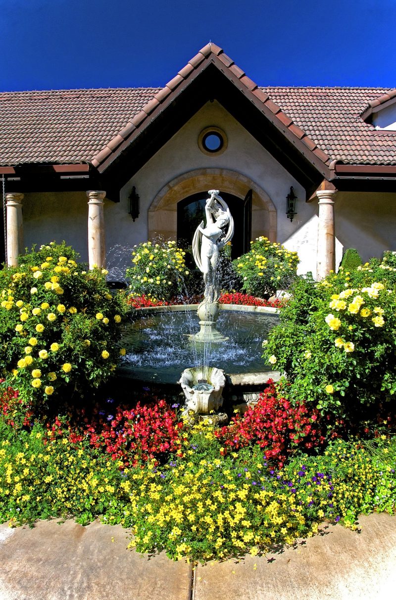 circular stone fountain with handcrafted siding and Italian dancer stone statue with clamshell spout spraying water into fountain and arched stone front entry with yellow rosebushes