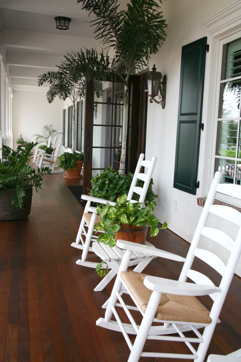 dark forest green wooden window shutters and white rockers with rush seats on stained hardwood floor with glass swing out double doors with wooden muntins and vintage barrel planters