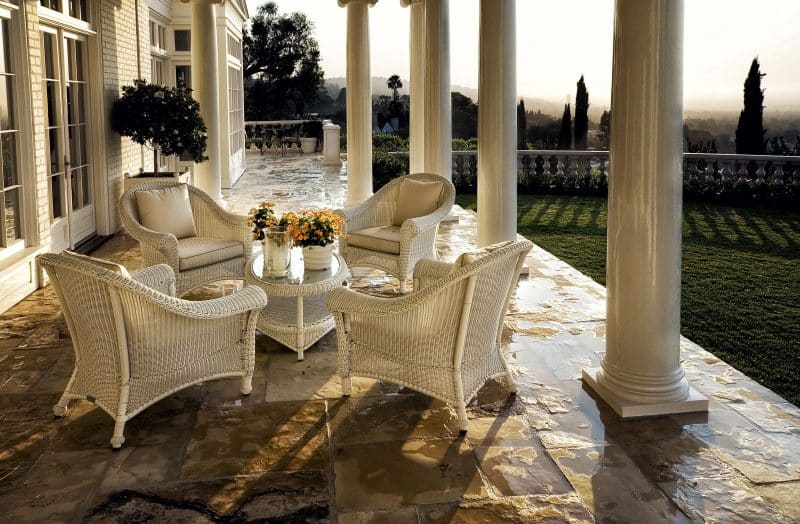 DIY box-shaped planter with wrap around porch with stone flooring and Roman ionic columns and white rattan chair and side table with ivory-hued seat cushions and pillows