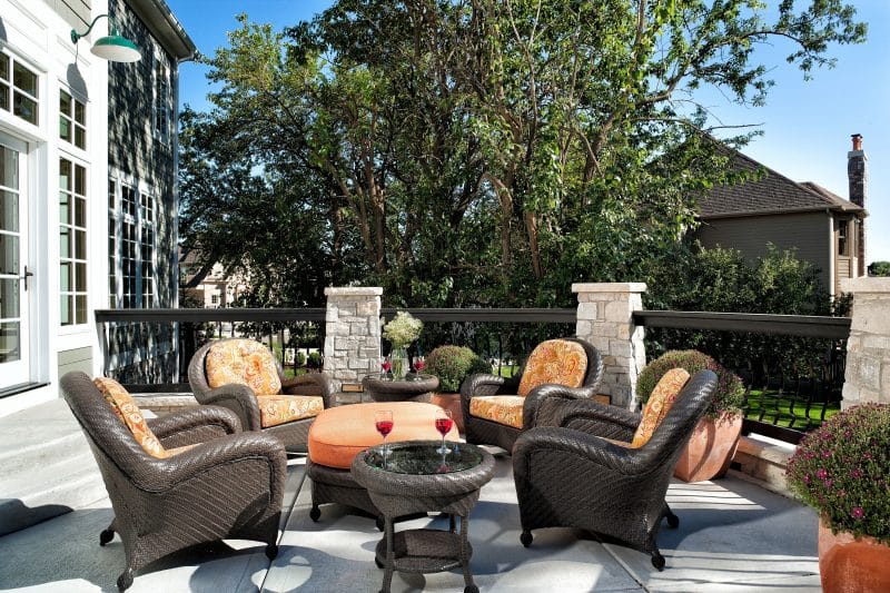 large tapered square terracotta planter with large round clay pots and synthetic resin wicker chairs and side tables with peach cushion seats with graphic print and half circle upper balcony and deck rail stone columns with iron railings with wavy balusters