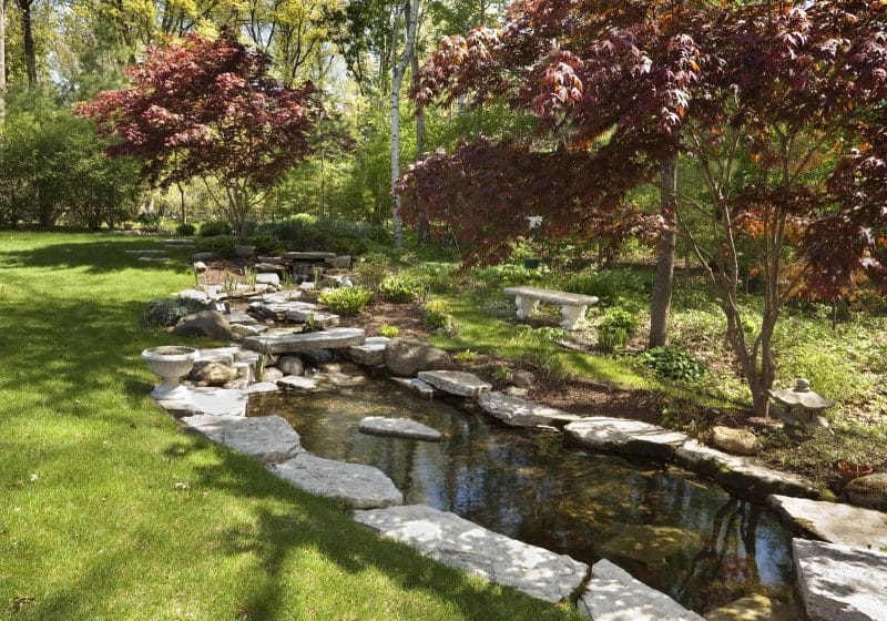 natural-narrow-stream-like-waterfall-pond-with-slab-pond-edges-and-stone-bench-and-white-stone-urn-planters-with-stone-slab-makeshift-bridge