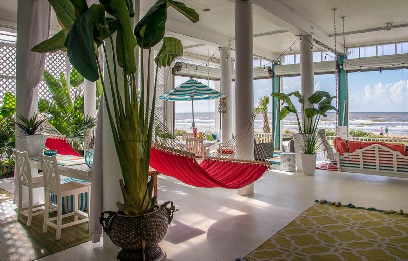 painted lattice porch siding with transom windows and red fabric hammock and wooden swing bench with motorized roll-up doors and string lights and glazed white tapered planters