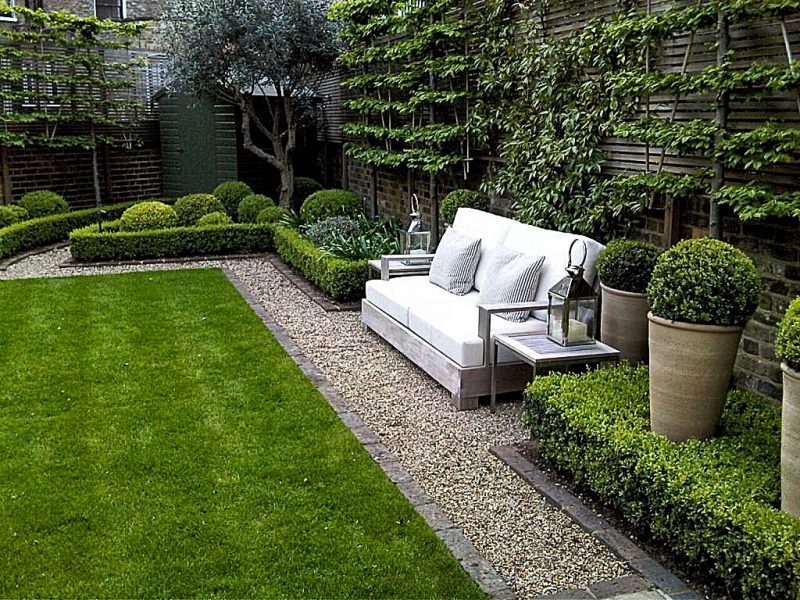 pea gravel walkway with brick garden edging and wooden bench with white cushions and thin pinstripe pillows and boxwood hedges and topiaries with citronella candle holder on side tables and trimmed topiaries in large clay planters