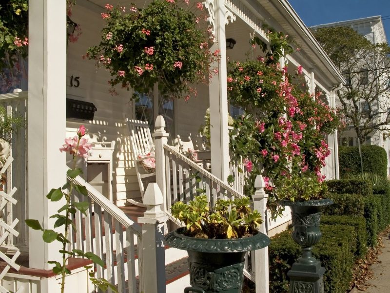porch spandrels with swirl brackets and pair of dark green urn planter and wooden balustrade with gothic newel caps and terra cotta painted porch floor two-tones with stairs and trimmed hedges and vintage mailbox