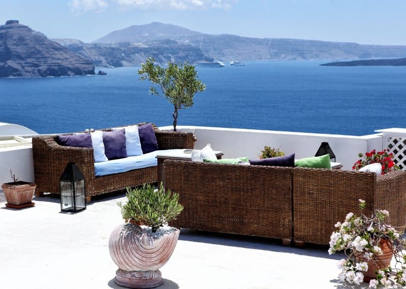 sea shell-inspired terra cotta planter with a white washed coating and olive tree with braided trunk and crushed white stones on top of planter with mature ivy geranium in terracotta olive jar blooms and Synthetic resin wicker sofa with seat cushion and colorful back pillows