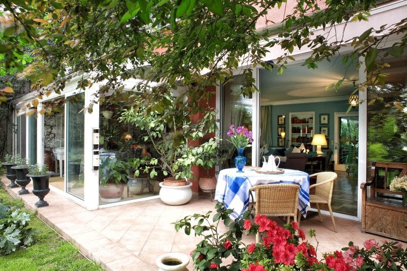 terracotta planter on copper stand with black Victorian urns outside sunroom and potted ficus tree placed on cast stone planter and rattan chairs with light pink patio tiles calling back terracotta planters