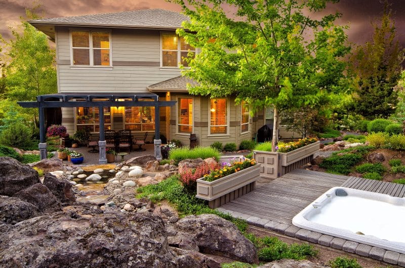 vibrant glazed blue planter calling back pergola rafters and natural stones and granite boulders with pond and twin flower box marking the entrance to jacuzzi deck with semi-hexagon sunroom with access to pergola and ornamental grasses