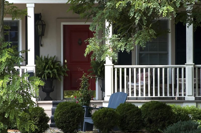white porch balustrade with raised panel on column bases and red front door with chrome kickplate and pair of antique cast iron urn planters with ferns and stair runner calling back the planters