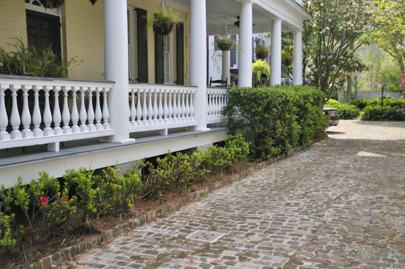 wraparound porch with light gray painted porch flooring calling back the balustrade top and olive green fixed shutters with weathered brick pavers and garden edging