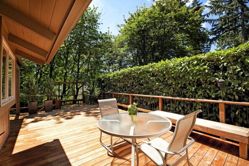 young pine trees on tall resin planters with au naturel clipped rivacy hedge and wooden deck planks and railings with custom made deck chairs with wavy backs and backless DIY wooden benches