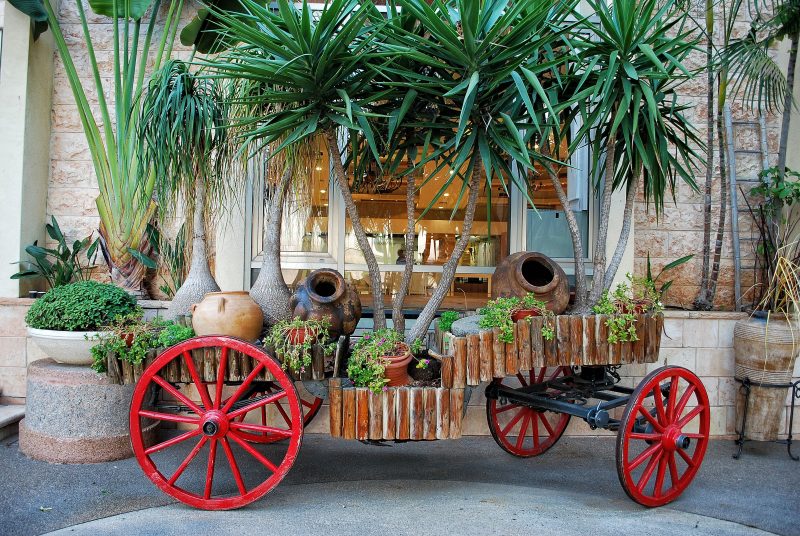 3-level log wagon garden bed with dark iron tires with contrasting red spokes and rim with grouped terracotta planters of different size and styles with antique Greek antique terra cotta jar on iron stand