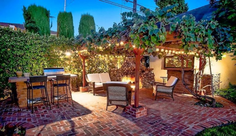 brick base for wooden pergola posts and stacked stone fireplace with stone veneer outdoor kitchen countertop base and 2 bar stools with wicker backrests with curved brick floor edging