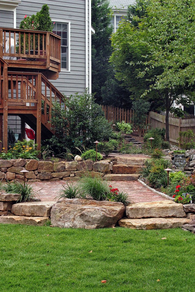 brick entryway with raised pond and waterfall edging and boulders and stone landscaping with dark step stones leading to backyard and cobblestone edging