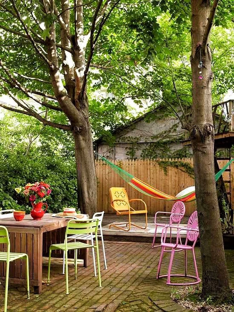 brick flooring in offset pattern with curved brick soldier course edging around tree and multicolored hammock with multicolored metal chairs and wooden table with slatted legs and bare wood French gothic picket fence