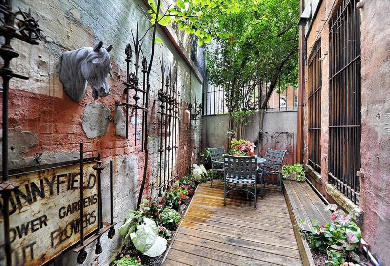concrete and wrought iron privacy fence with rough concrete wall with antique bronze wrought iron grilles and salvaged flower shop sign with 3 gray basketweave steel chairs and raised garden bed with built-in bench