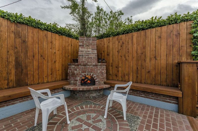 corner brick arched fireplace with raised brick hearth and 2 antique bronze lanterns on fireplace mantel with brick and exposed aggregate concrete medallion inlay on floor and wood plank fencing with built-in benches