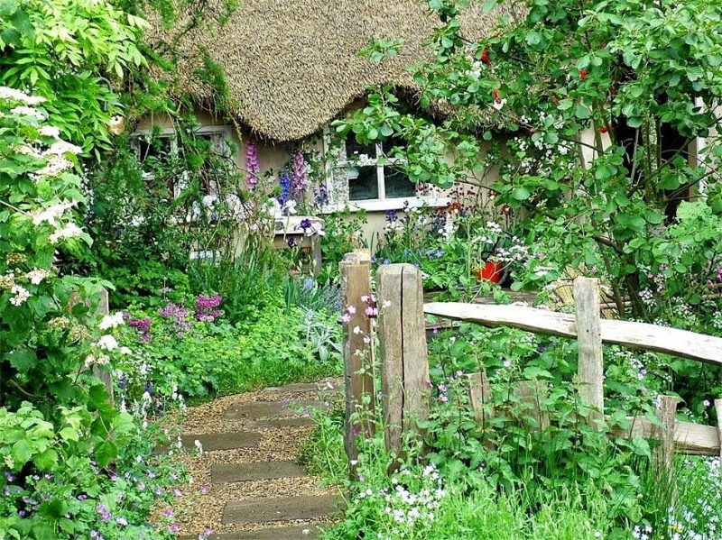 cottage with thatched roof with split rail fence and pea gravel walkway with stepping stones and white and pastel flower blooms on walkway and facade with strawberry guava tree