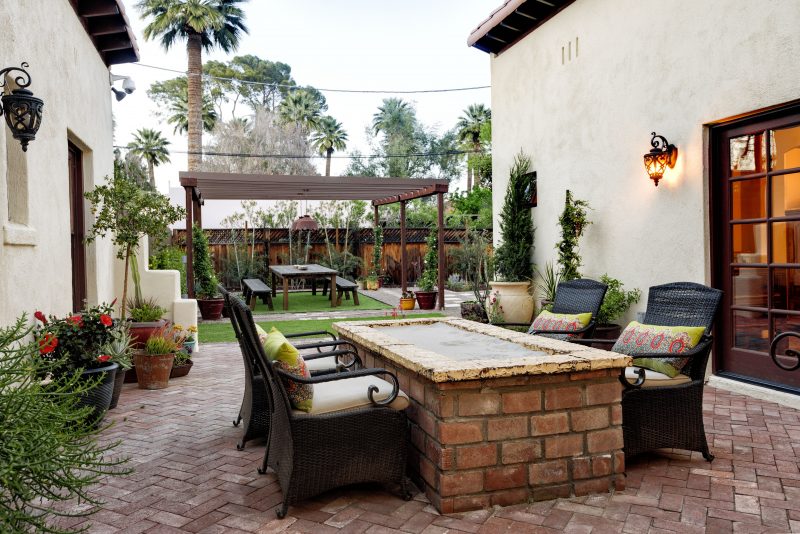 gas fire pit with sand fill and running bond brick base with herringbone brick flooring and black wicker chairs with scrolled armrests and oil-rubbed bronze wall sconces with brown pergola over matching table and benches