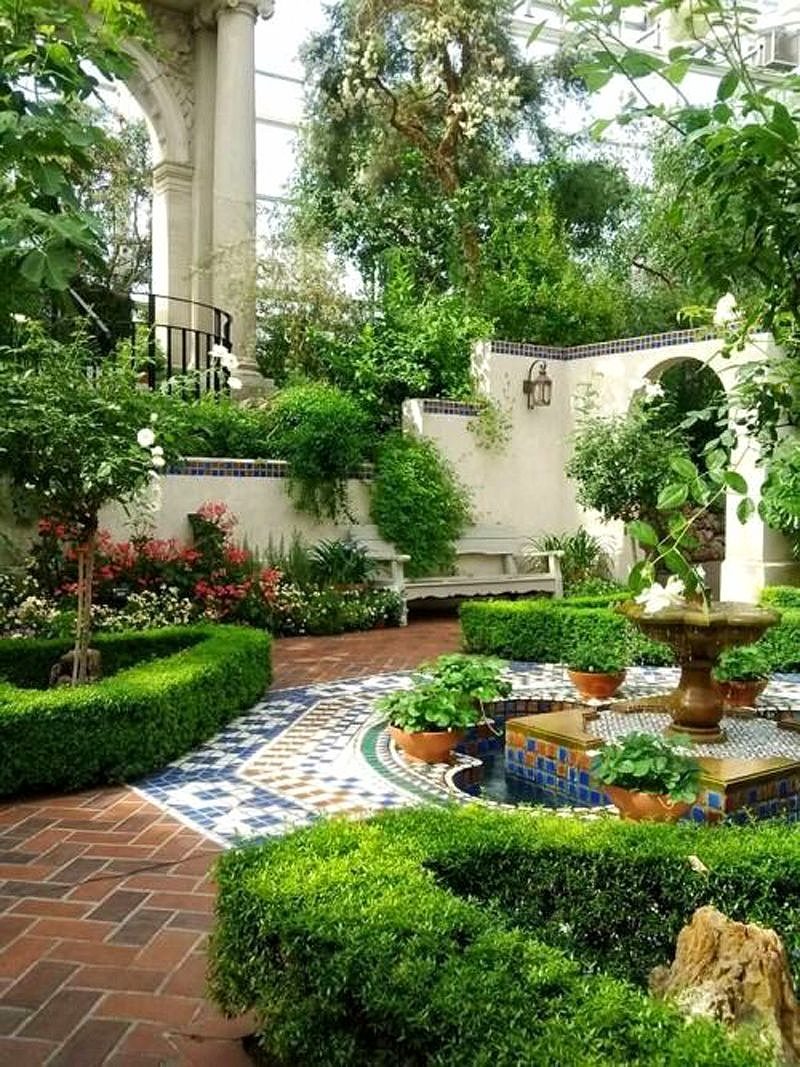 herringbone brick flooring with multicolored porcelain inlay and flower-shaped water fountain with sculpted shrubs and white wooden bench against white wall with blue porcelain mosaic tile trim