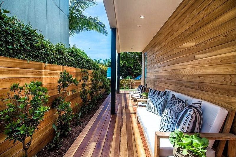 horizontal cedar fence with built-in planter on top and unfinished wooden sofa with white cushions and black and white patterned throw pillows with wood planked covered narrow deck and contrasting black deck roof posts