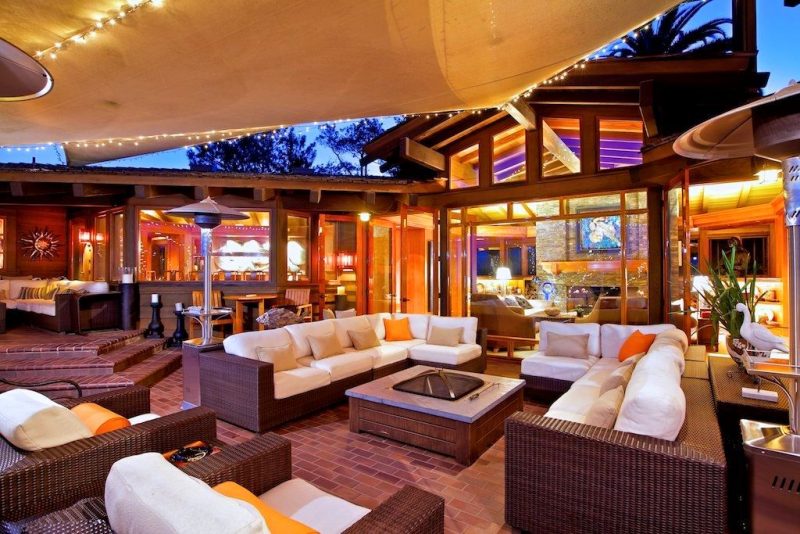 multi-toned brown brick floor and soapstone topped square gas fire pit with outdoor wicker seating furniture with white cushions and orange throw pillows and 3 outdoor patio heaters with beige canopy with string lights