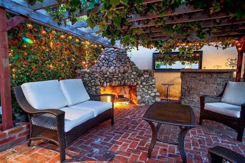 red brick flooring with stacked stone open fireplace and brown wicker chairs with matching tiered table and natural rattan ball string lights with black framed rectangular mirror and wooden pergola with climbing vines