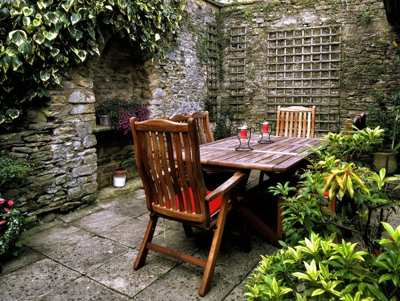 stacked natural stone walls with duo bamboo trellises arched alcove for planters with stone shelf and wood planked table with slatted wood back dining chairs and weathered concrete tiled floor