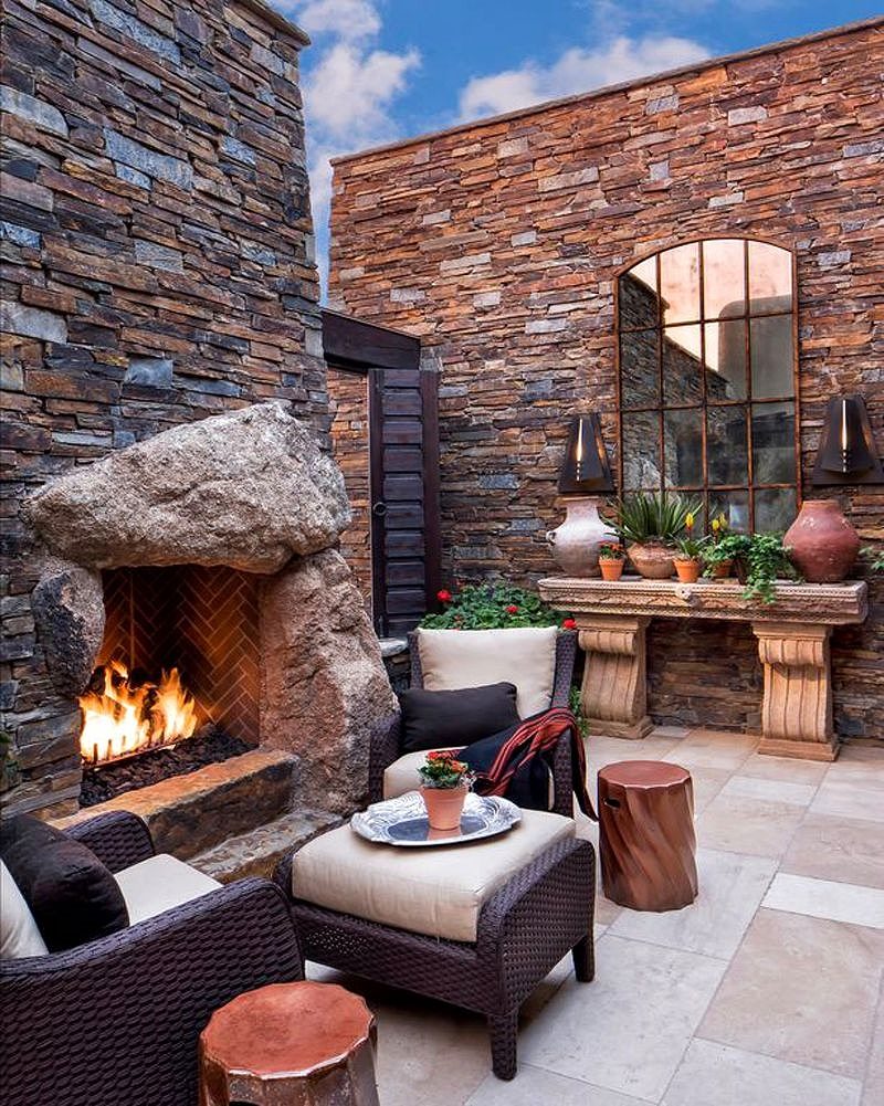 stone veneer patio walls and segmental arched window mirror with open fireplace with boulder capstone and herringbone tiled firebox with cast stone console table with multi-sized planters and 2 copper spiral fluted end tables