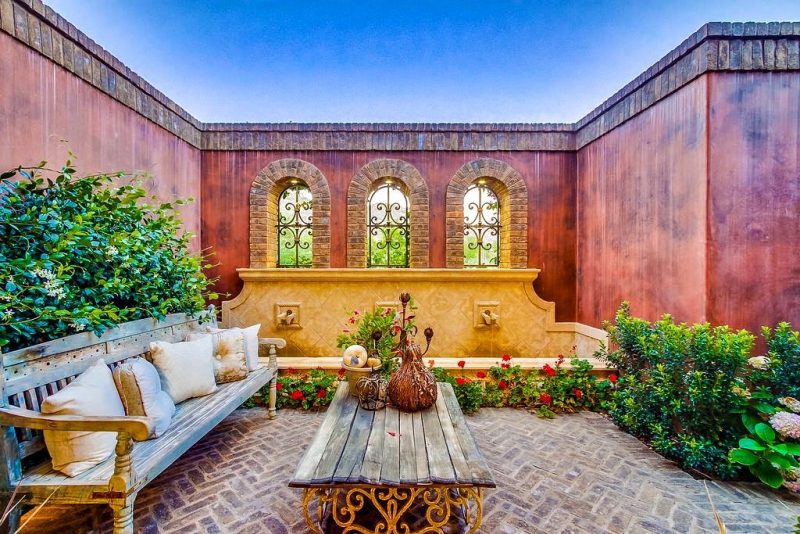 weathered brick patio floor and 3 arched brick framed windows with wrought iron grilles and reclaimed planked coffee table with yellow wrought iron legs and milk painted long bench with throw pillows