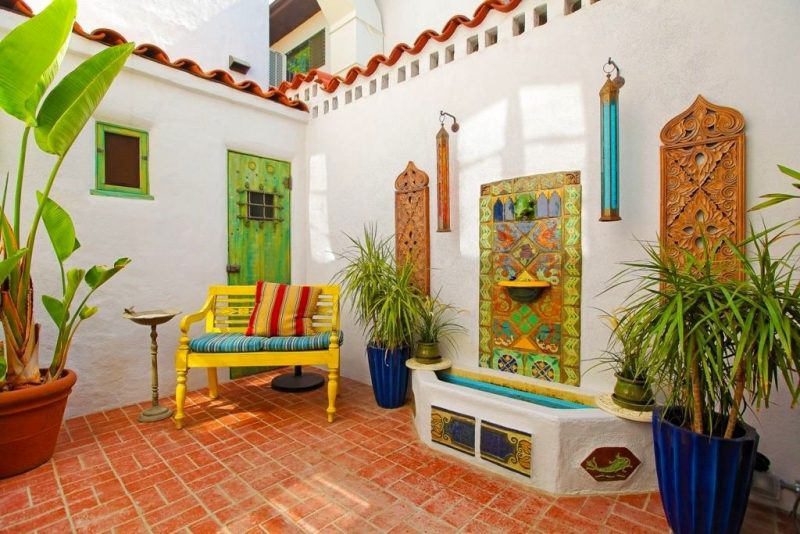 white walls with 2 Moorish wood wall panels and handpainted terracotta wall tiles with yellow semi-circle shelf with yellow wooden settee and 2 blue fluted planters with 2 colored glass and brass cylinder wall sconces