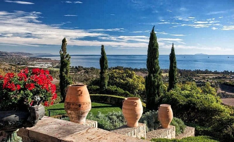 antique bronze low urn planter with patina and three ridged terracotta olive jars with stone staggered retaining wall with boxwood hedges and tall evergreen trees