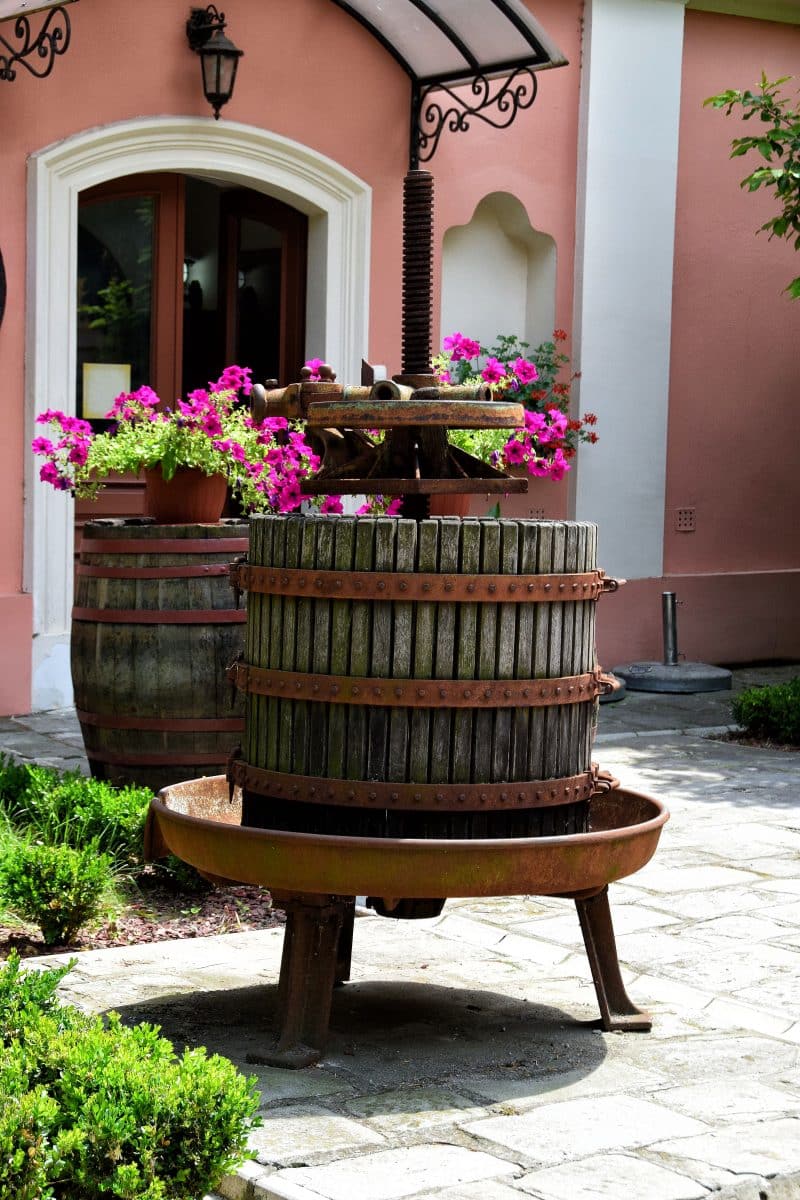 antique wood and cast iron base grape press and hogshead barrel used as plant riser with pink petunias and arched doorway with pink stucco siding and polycarbonate door canopy with swirl brackets