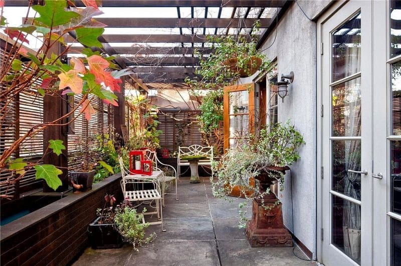 bronze bird bath in a rust patina repurposed as planter with attached pergola with privacy walls and round table with moss-covered top and wooden sliding louvre windows with wall mounted marine lantern style patio lighting
