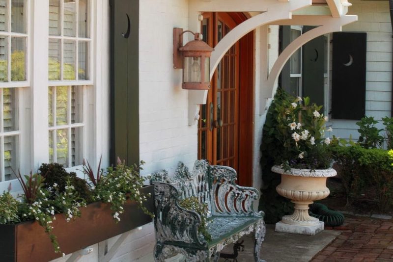 cast stone pedestal planter with white azaleas and rectangular window box planter with antique iron settee with verdigris and arched country French front door with dark gray window shutters with crescent moon cutouts