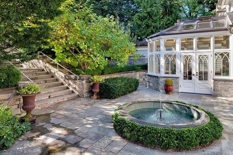 pair of antique cast iron urn planters and circular pond with water fountain and slate tile pond coping with stacked stone retaining wall and sunroom with stone base and boxwood hedges covering perimeter of pond