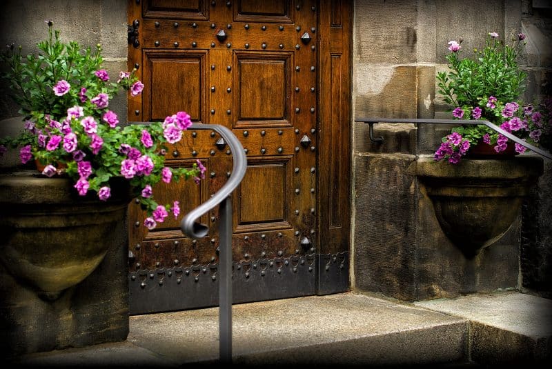 pair of weathered built-in wall planters with pink petunias and custom recessed panel wooden front door with iron rivets and iron kick plate