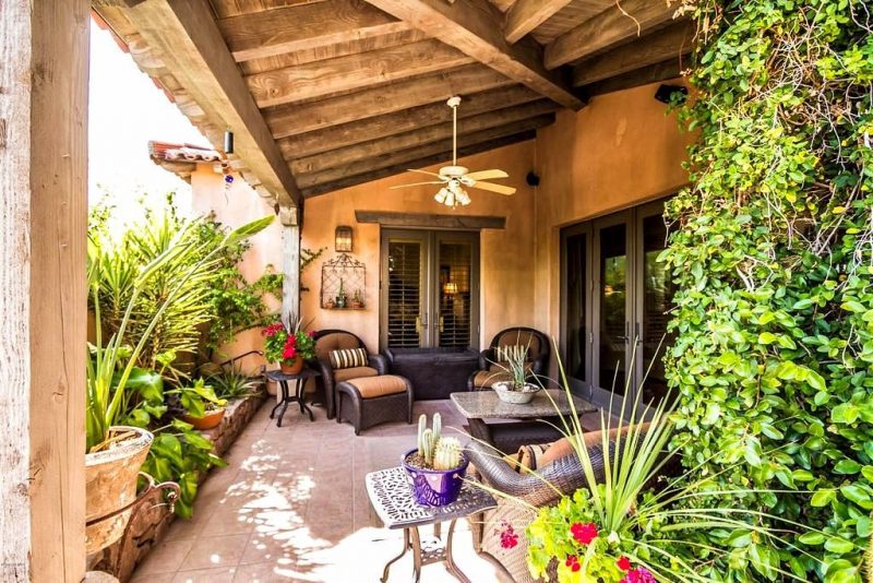 reclaimed beams on wood planked patio ceiling with 2 black iron lattice side tables and wrought iron wall-mounted planter holder and 3 armchairs with matching foot stools and travertine tile flooring